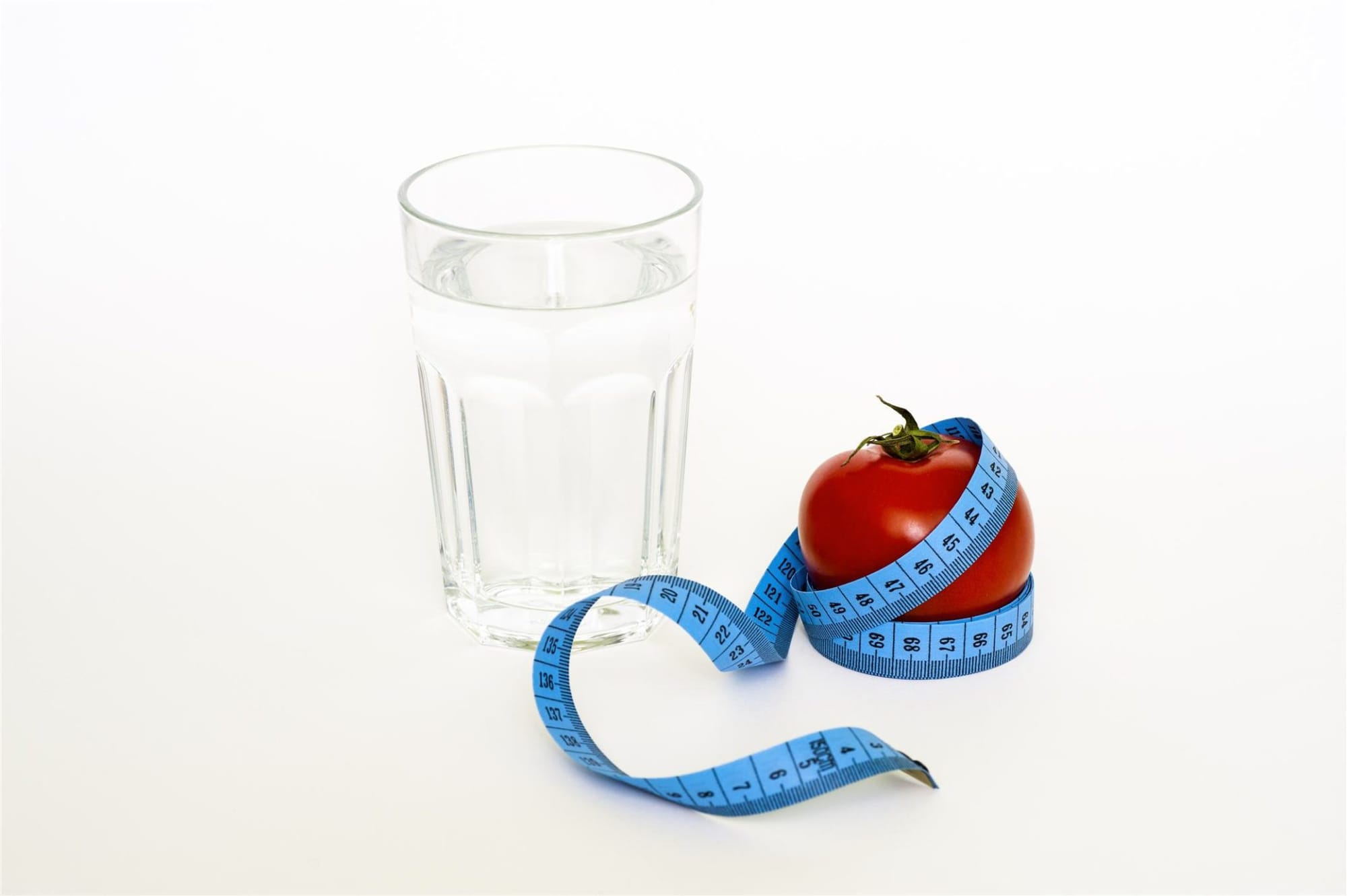 A glass of water and a tomato wrapped in a blue measuring tape, symbolizing weight loss and healthy eating