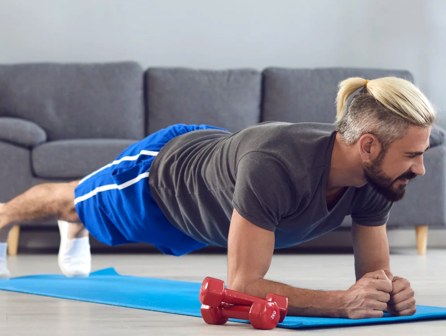 Man with a blonde ponytail holding a forearm plank on a blue mat, with red dumbbells in front of him in a living room.