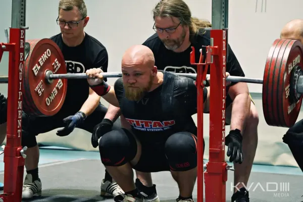 Powerlifter squatting with a loaded Eleiko barbell, supported by spotters during a competition