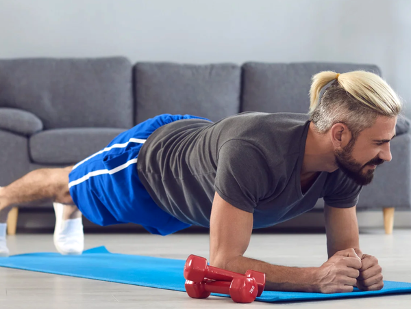 Man with a blonde ponytail holding a forearm plank on a blue mat, with red dumbbells in front of him in a living room.