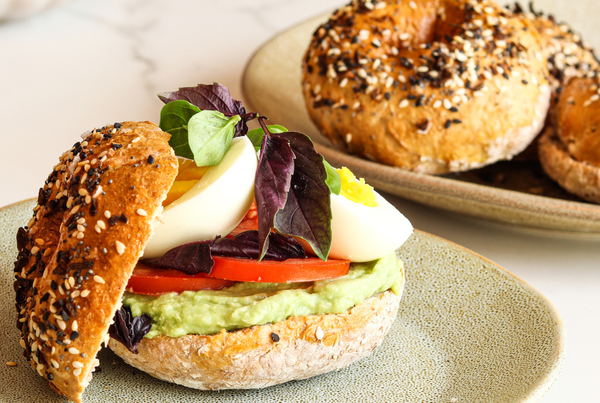 High-protein bagel with avocado, tomato, boiled egg, and fresh greens on a plate, with extra bagels in the background.