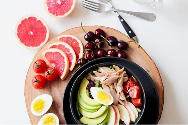 Balanced meal with avocado, chicken, eggs, tomatoes, and greens in a bowl, surrounded by grapefruit, cherries, and eggs